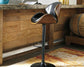 The Bellatier Tall UPH Swivel Barstool features adjustable height, a black metal base, wood accents, and a black faux leather cushioned seat, shown at a rustic wood bar counter with a wine barrel beside it.