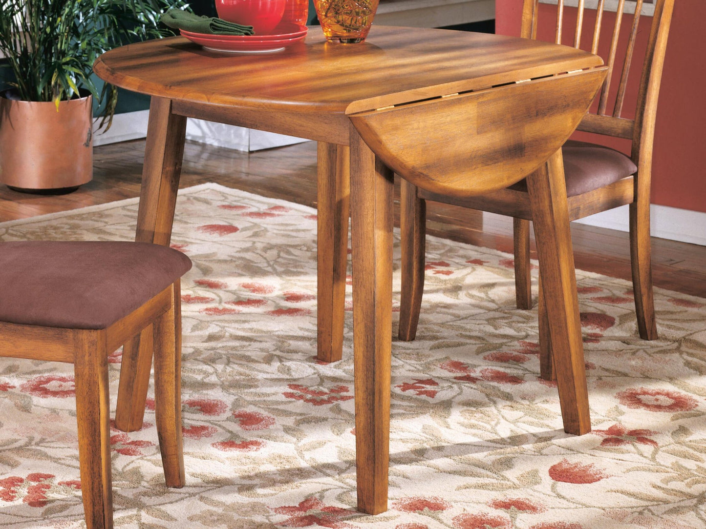 The Berringer Round DRM Drop Leaf Table with two chairs stands on a floral rug, set with dishes in a sunlit room.