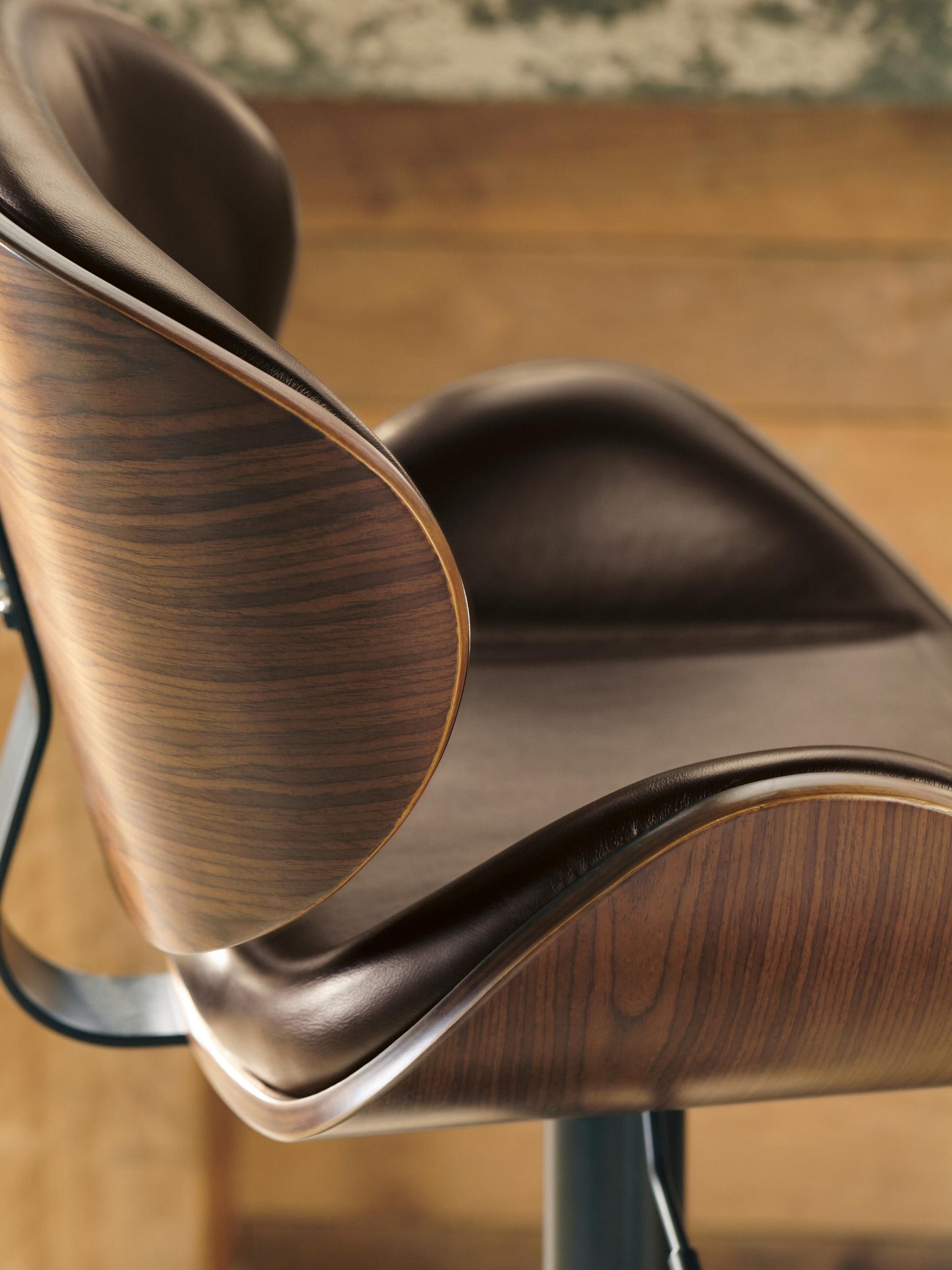 Close-up side view of the Bellatier Tall UPH Swivel Barstool, featuring a curved wood veneer backrest and brown faux leather upholstery, set against a wooden background.