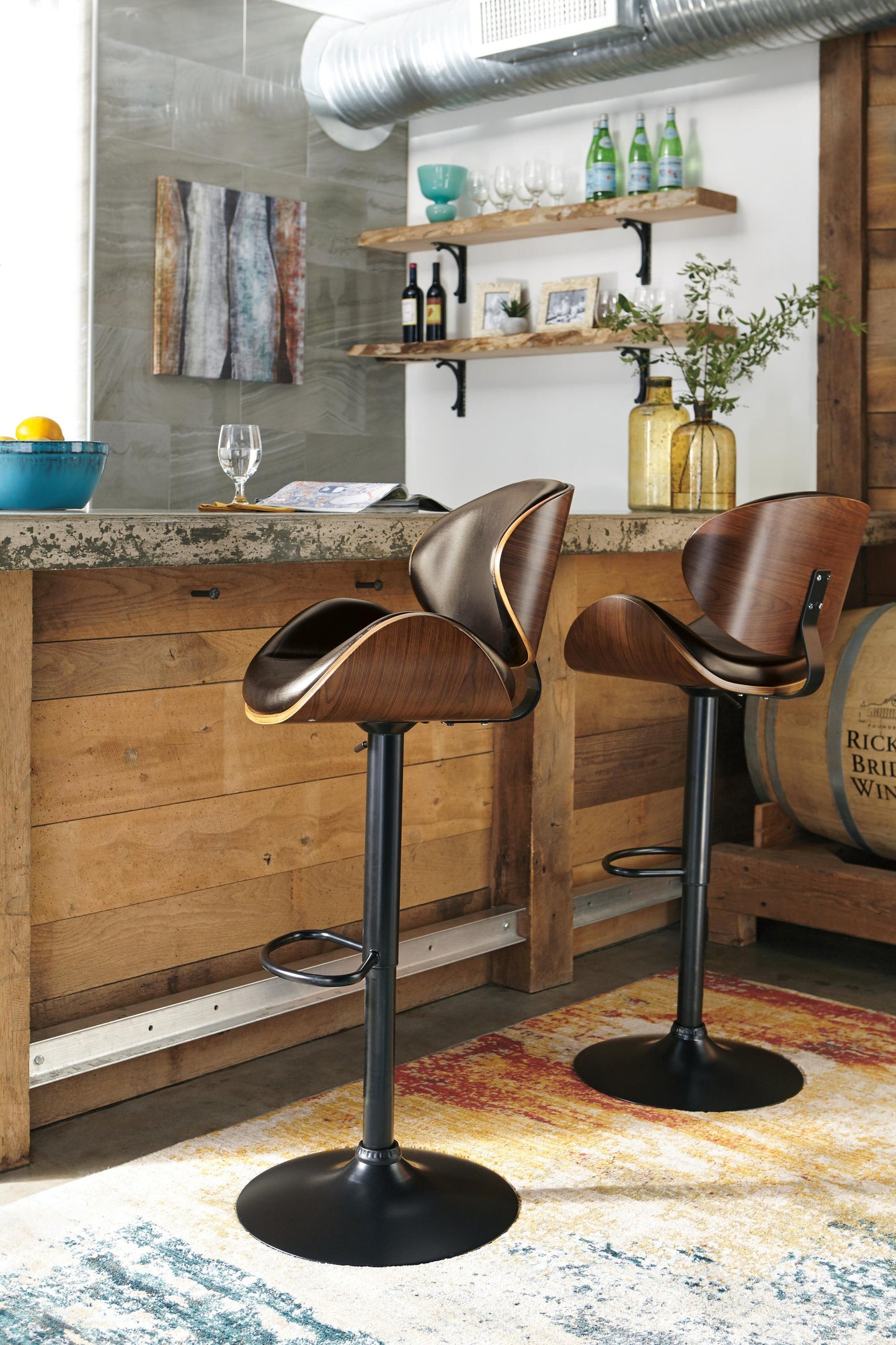 A pair of Bellatier Tall UPH Swivel Barstools with adjustable height and faux leather upholstery are placed at a rustic kitchen counter, with shelves of glassware and bottles behind them and a colorful rug on the floor.