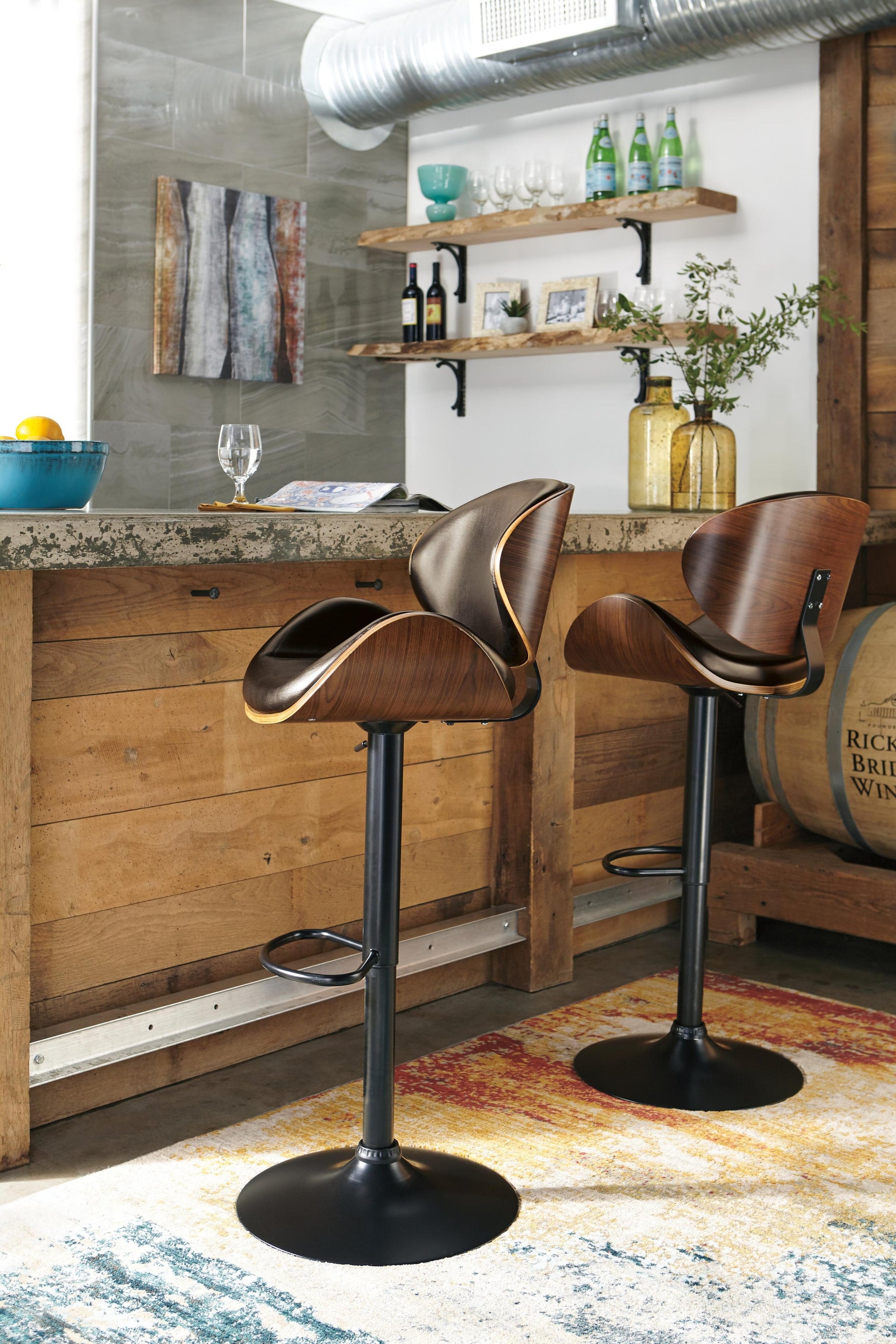A pair of Bellatier Tall UPH Swivel Barstools with adjustable height and faux leather upholstery are placed at a rustic kitchen counter, with shelves of glassware and bottles behind them and a colorful rug on the floor.