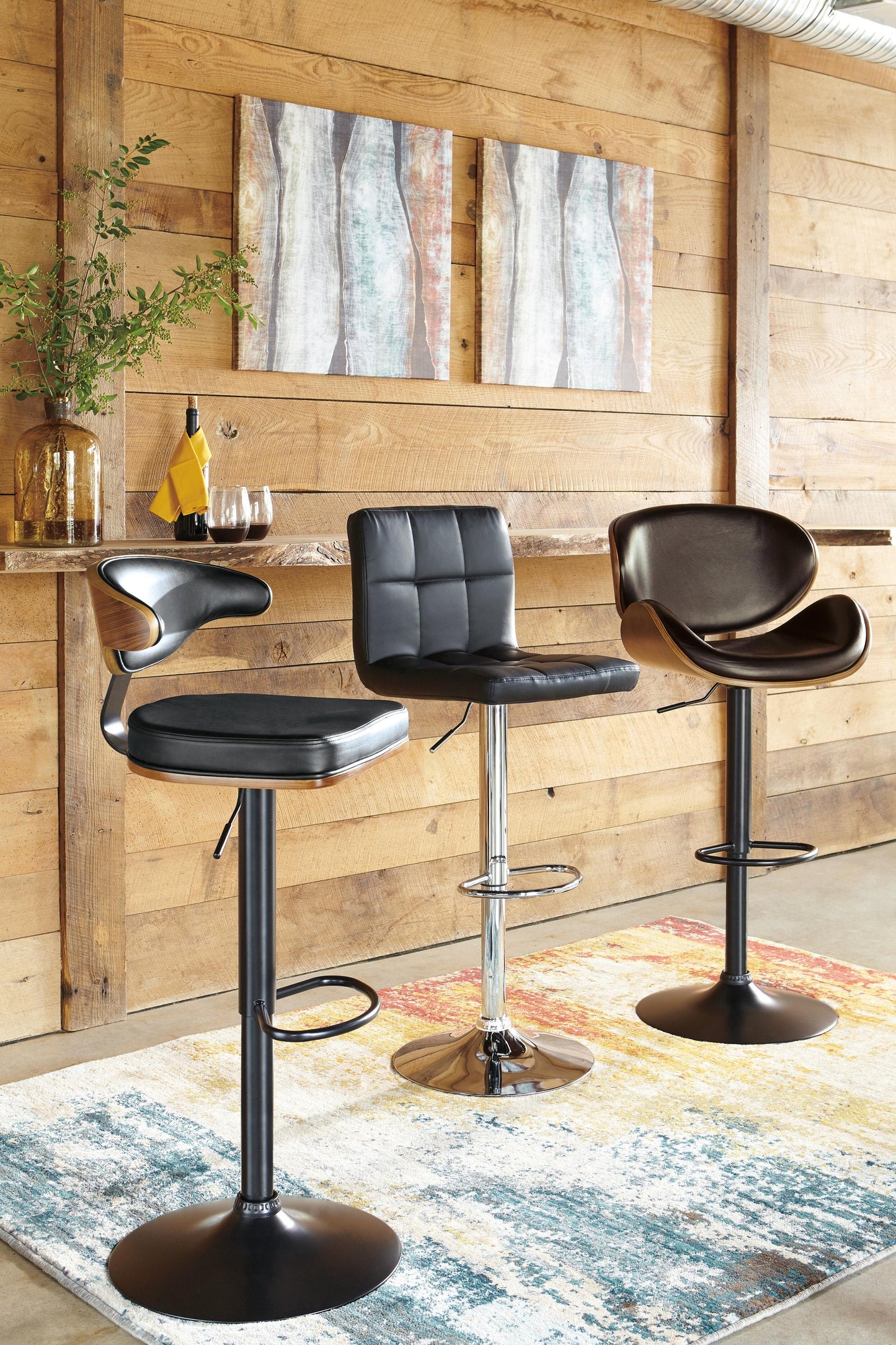 Three Bellatier Tall UPH Swivel Barstools with black and brown faux leather upholstery are arranged at a wooden bar counter in a room featuring wood-paneled walls, wall art, and a colorful rug.