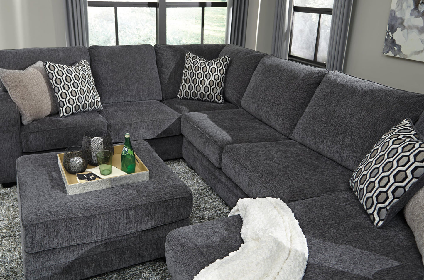 A contemporary gray sectional sofa with patterned pillows and a white blanket pairs with the Tracling Oversized Accent Ottoman, topped with a tray, glasses, and bottle, all in a living room with large windows and slate gray fabric upholstery.