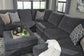 A contemporary gray sectional sofa with patterned pillows and a white blanket pairs with the Tracling Oversized Accent Ottoman, topped with a tray, glasses, and bottle, all in a living room with large windows and slate gray fabric upholstery.