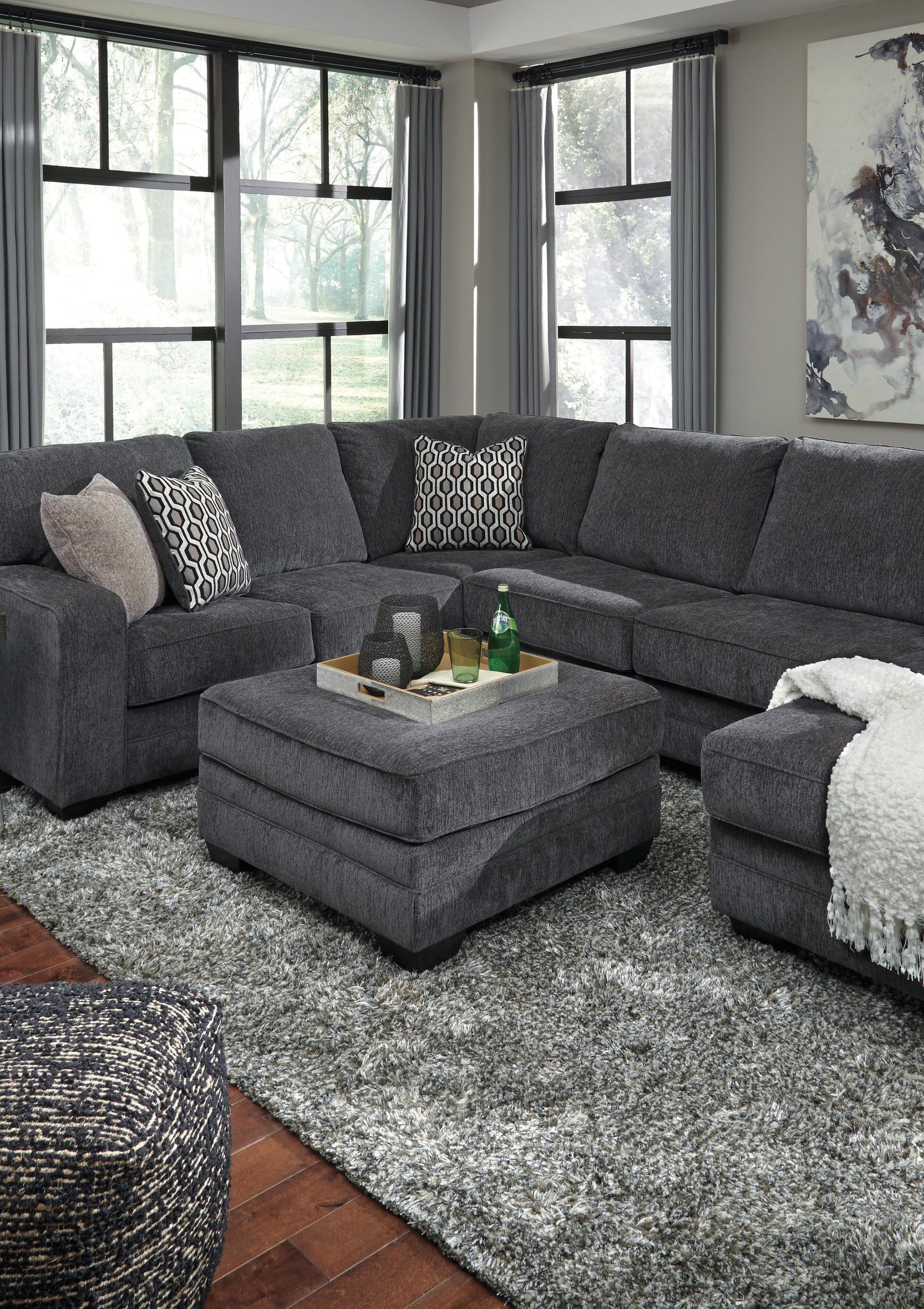 A modern living room features a slate gray fabric sectional, the Tracling Oversized Accent Ottoman, decorative pillows, a throw blanket, and a tray with drinks on a textured gray rug by large windows in contemporary style.