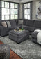A modern living room features a slate gray fabric sectional, the Tracling Oversized Accent Ottoman, decorative pillows, a throw blanket, and a tray with drinks on a textured gray rug by large windows in contemporary style.