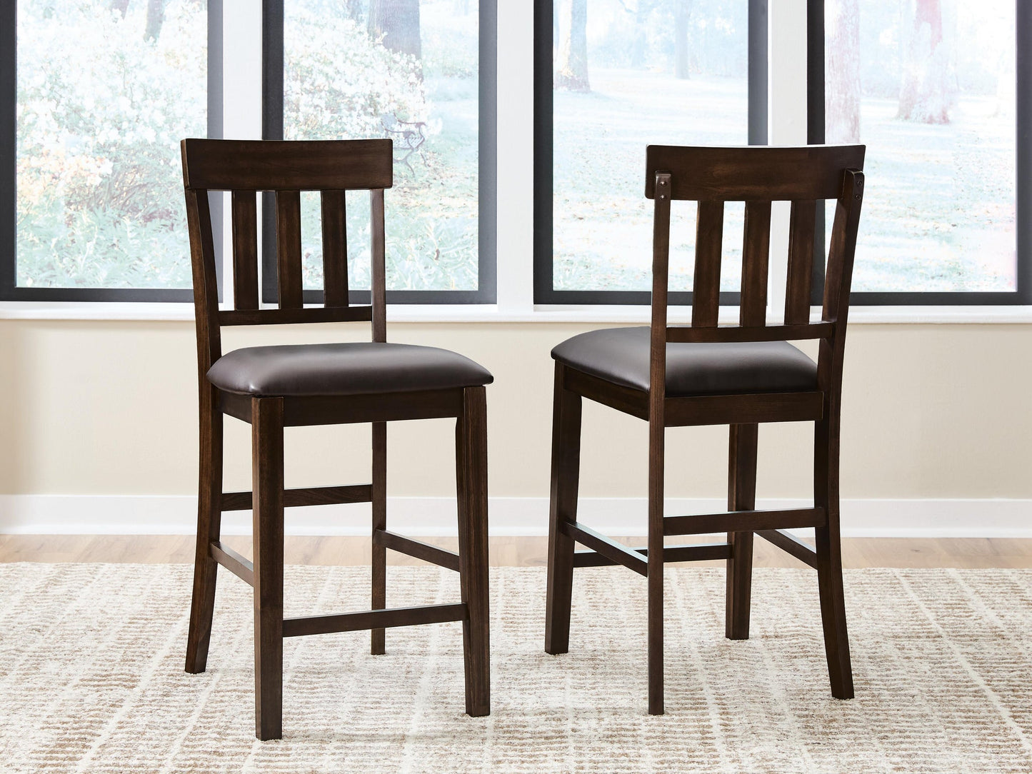 Two Haddigan Upholstered Barstools with dark wood, slat backs, and cushioned seats sit on a light patterned rug in front of large windows.