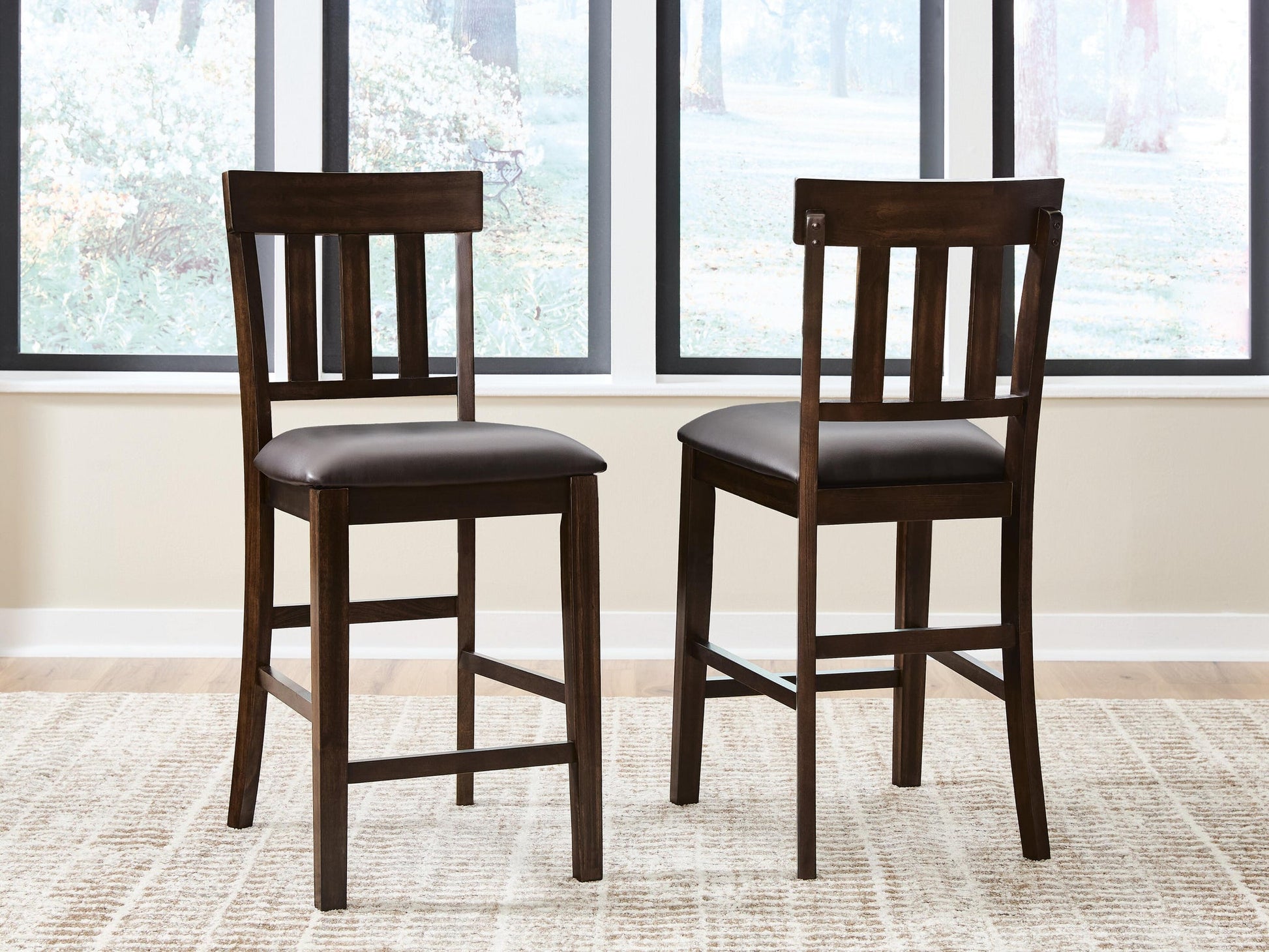 Two Haddigan Upholstered Barstools with dark wood, slat backs, and cushioned seats sit on a light patterned rug in front of large windows.