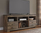 A flat-screen TV sits on the Trinell LG TV Stand w/Fireplace Option, featuring open shelves, drawers, and decor items; plaid curtains hang to the right.
