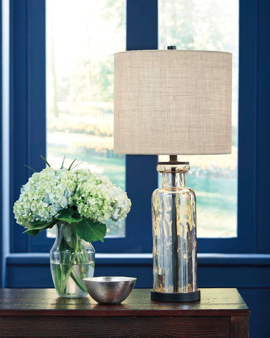 A Laurentia Glass Table Lamp, a glass vase with white hydrangeas, and a silver bowl sit on a dark wooden table in front of blue-framed windows.