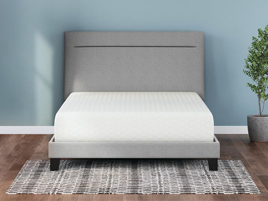 A neatly made bed with the Chime 12 Inch Memory Foam Mattress and a light gray upholstered headboard sits on a black-and-white patterned rug in a blue-walled room with a small potted plant.