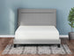 A neatly made bed with the Chime 12 Inch Memory Foam Mattress and a light gray upholstered headboard sits on a black-and-white patterned rug in a blue-walled room with a small potted plant.