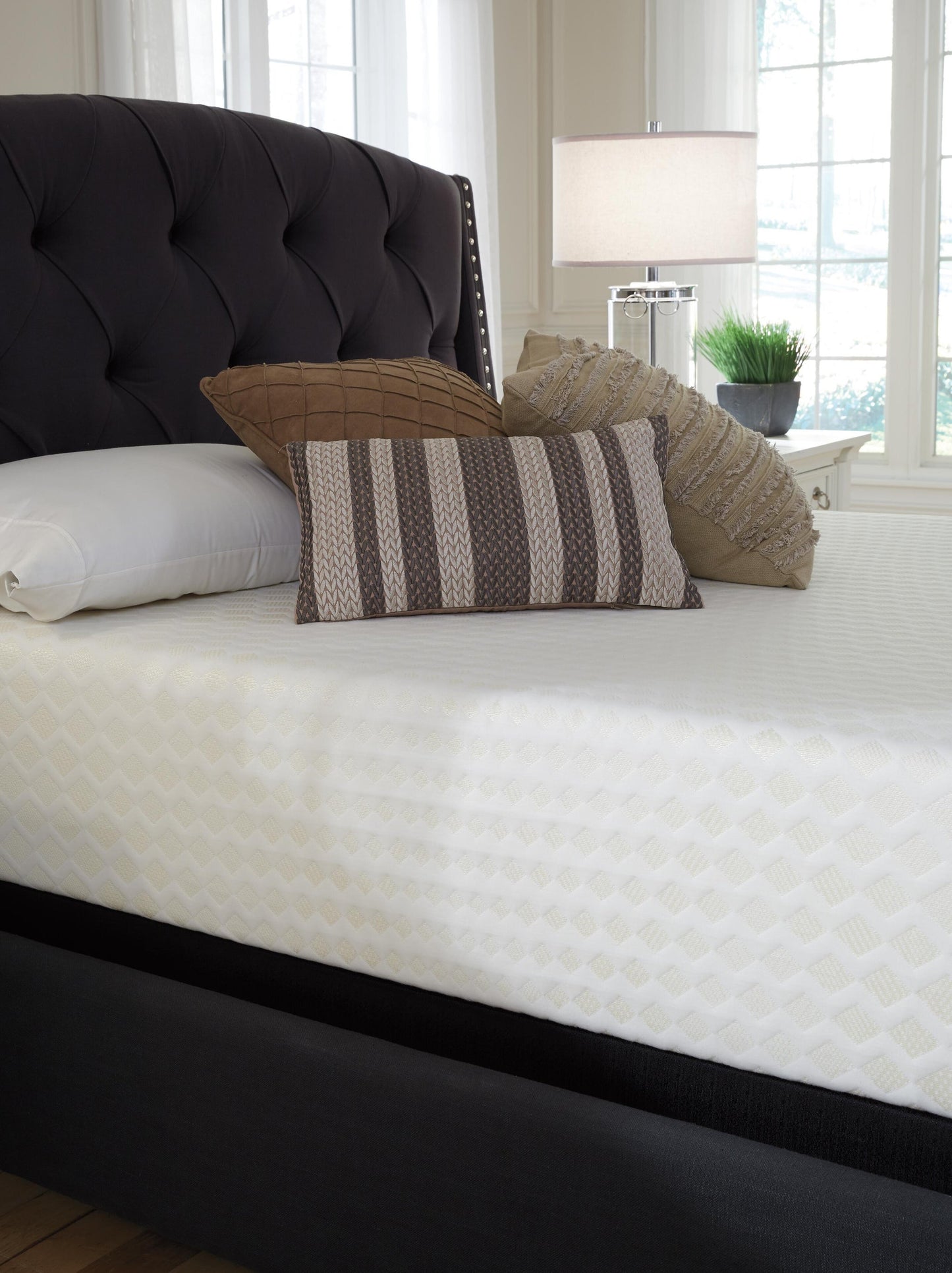A neatly made bed features a Chime 12 Inch Memory Foam Mattress (queen, white patterned) in a box, three decorative pillows, a headboard, and a bedside table with a lamp and potted plant.