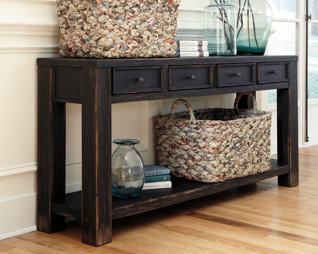 The Gavelston Sofa Table has a weathered black finish, four small drawers, and is styled with woven baskets, glass vases, and books on and below it. It sits against a white wall on a wooden floor.