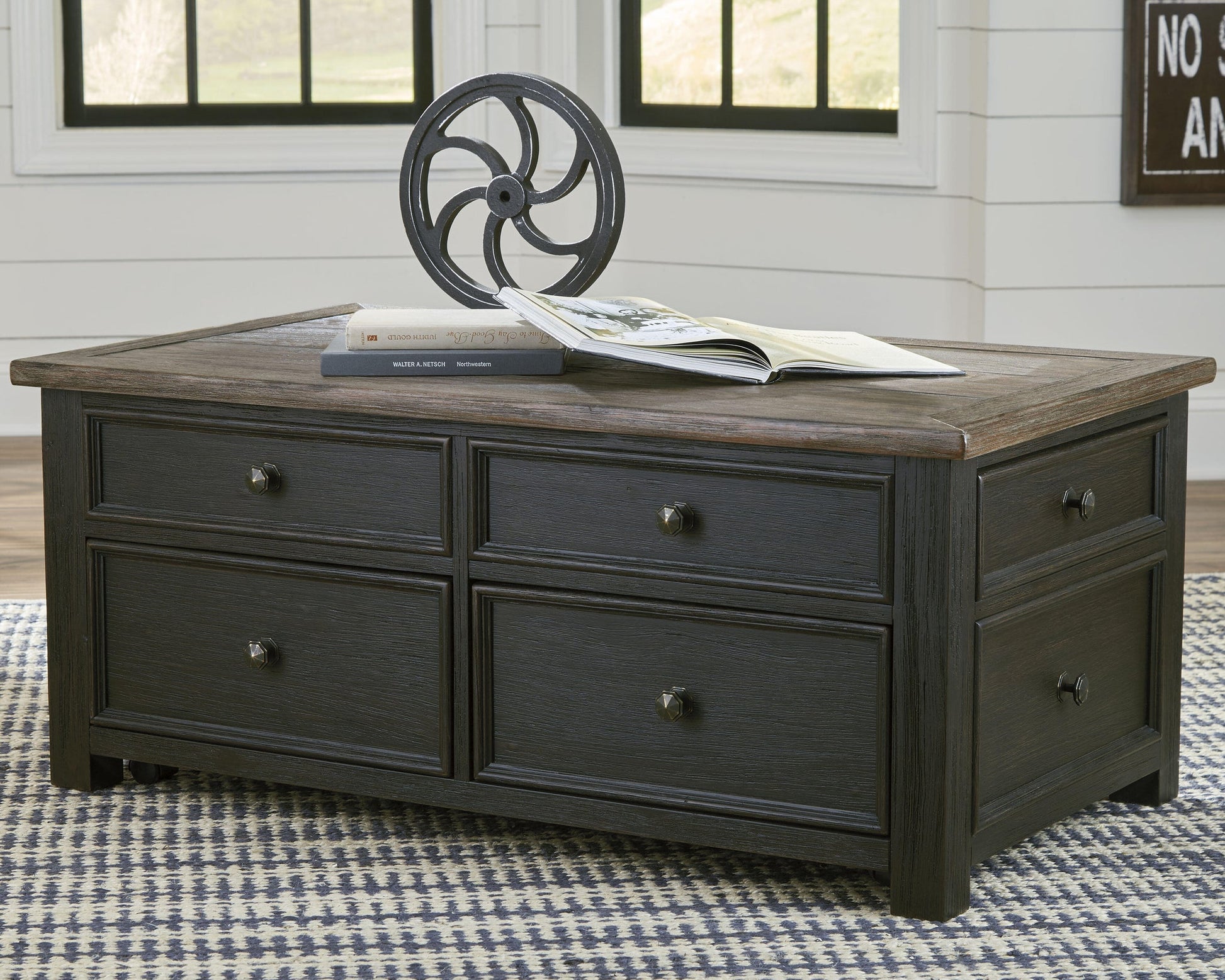 The Tyler Creek Lift Top Cocktail Table, featuring a dark wood finish and four drawers, is topped with a metal decorative object and two books, and placed on a patterned rug in a bright room.