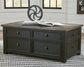 The Tyler Creek Lift Top Cocktail Table, featuring a dark wood finish and four drawers, is topped with a metal decorative object and two books, and placed on a patterned rug in a bright room.