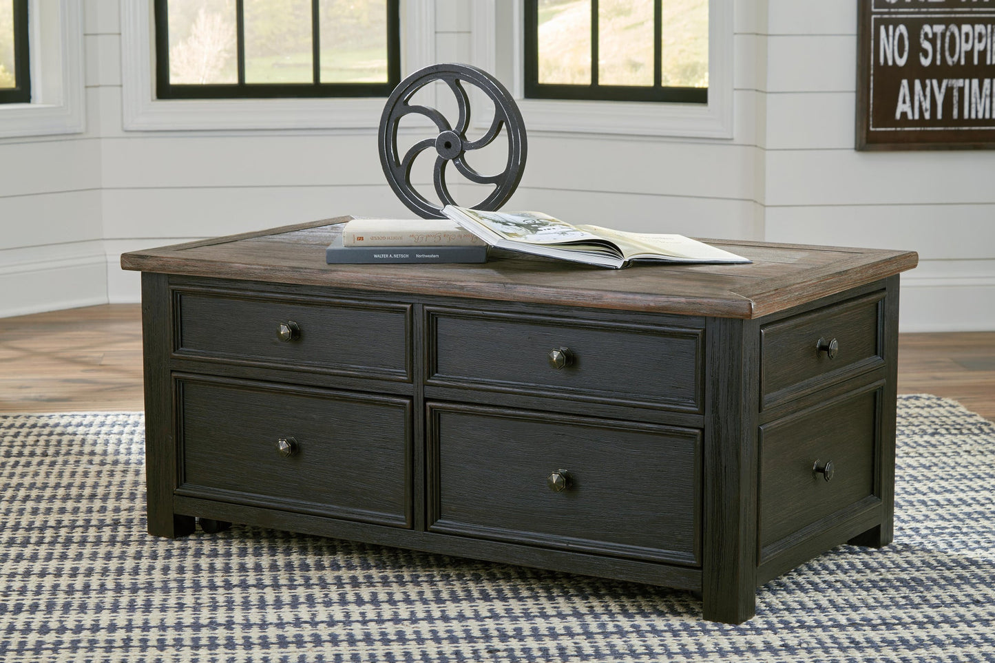 The Tyler Creek Lift Top Cocktail Table, made of dark wood with four drawers, displays a book and a decorative metal wheel on top and sits on a patterned rug in a bright room.