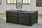 The Tyler Creek Lift Top Cocktail Table, made of dark wood with four drawers, displays a book and a decorative metal wheel on top and sits on a patterned rug in a bright room.