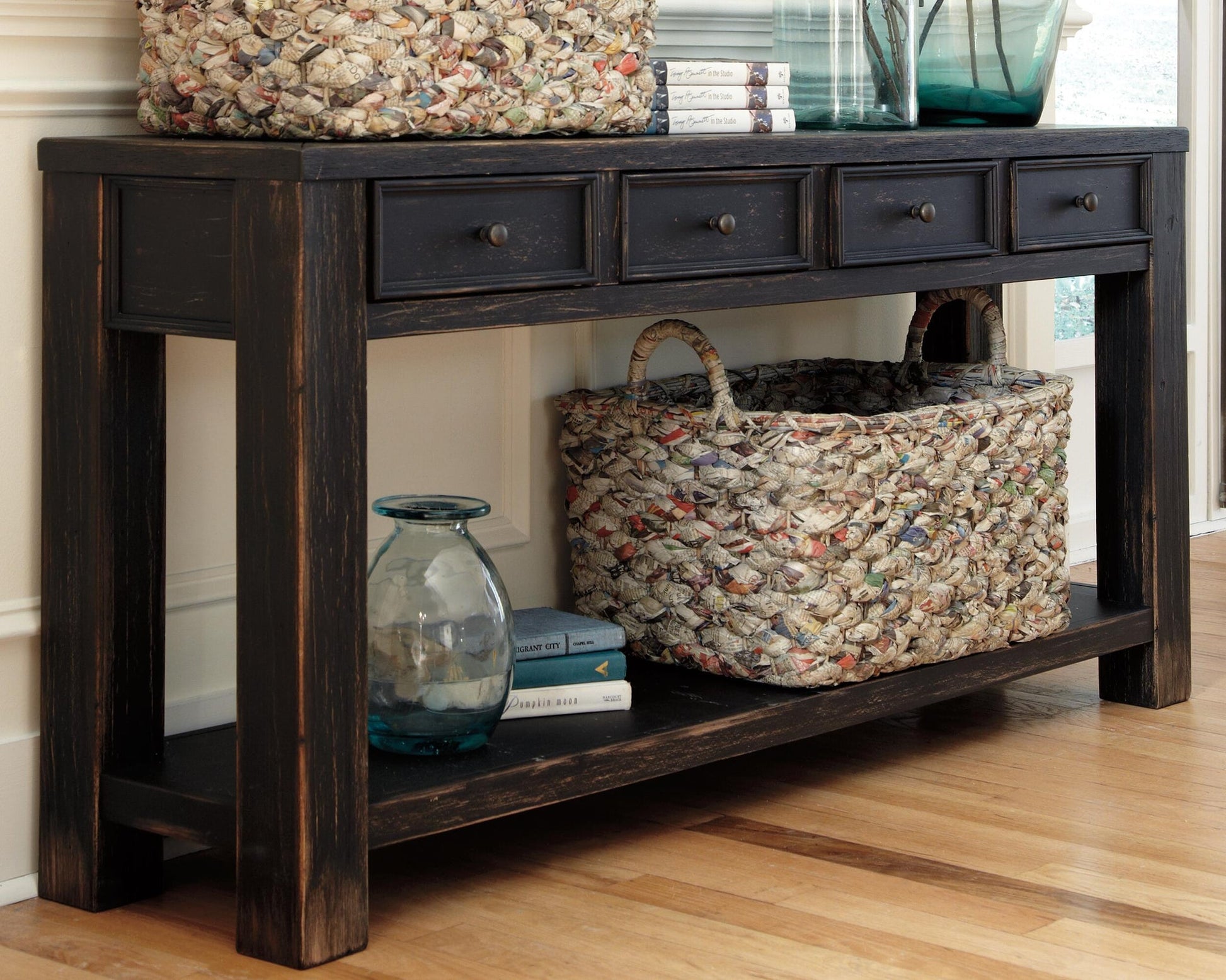 The Gavelston Sofa Table features a dark weathered black finish, four drawers, woven baskets on top and bottom, glass vases, books, and sits on a wooden floor.