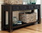 The Gavelston Sofa Table features a dark weathered black finish, four drawers, woven baskets on top and bottom, glass vases, books, and sits on a wooden floor.