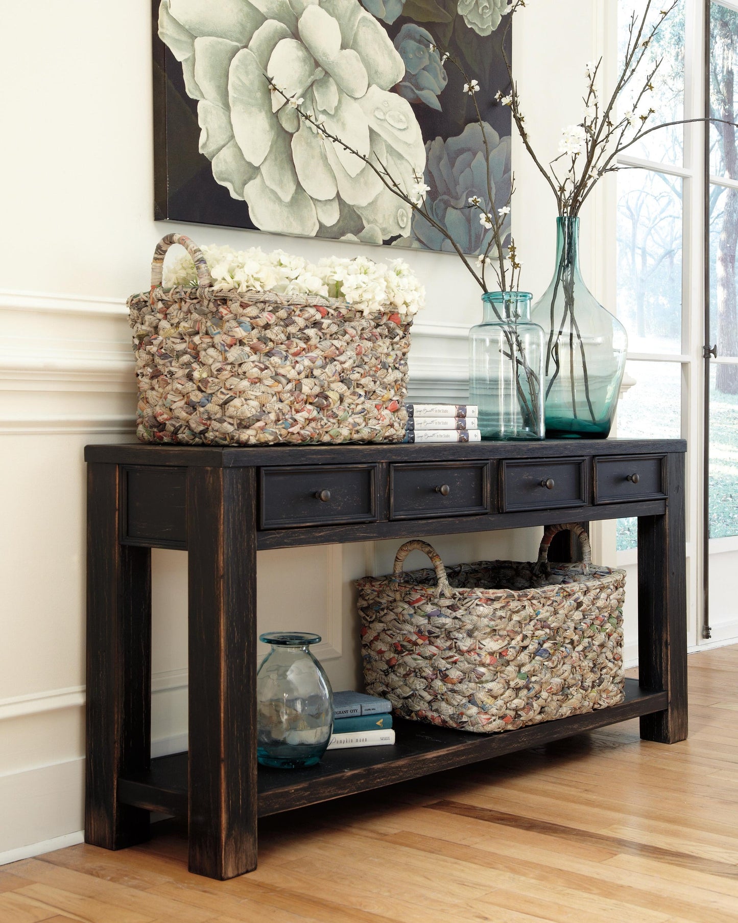 The Gavelston Sofa Table, with its weathered black finish, offers four drawers and two woven baskets for storage and is styled with glass vases, branches, and books beneath a bold floral painting for primitive charm.
