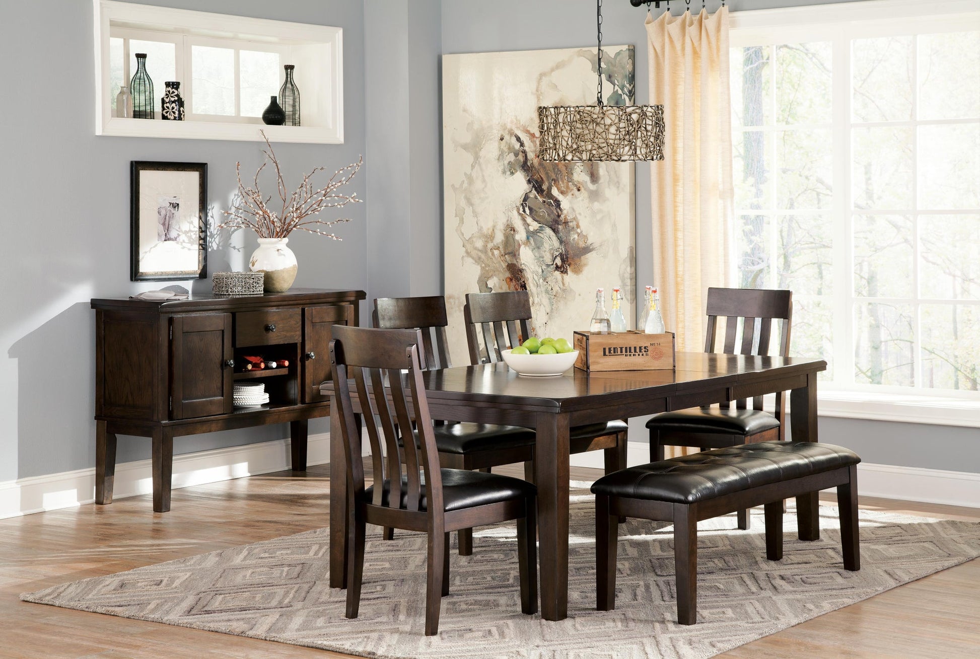 A dining room features a dark wood table, four chairs, the Haddigan Large UPH Dining Room Bench, a sideboard, modern decor, and a patterned rug beneath large windows with neutral walls.
