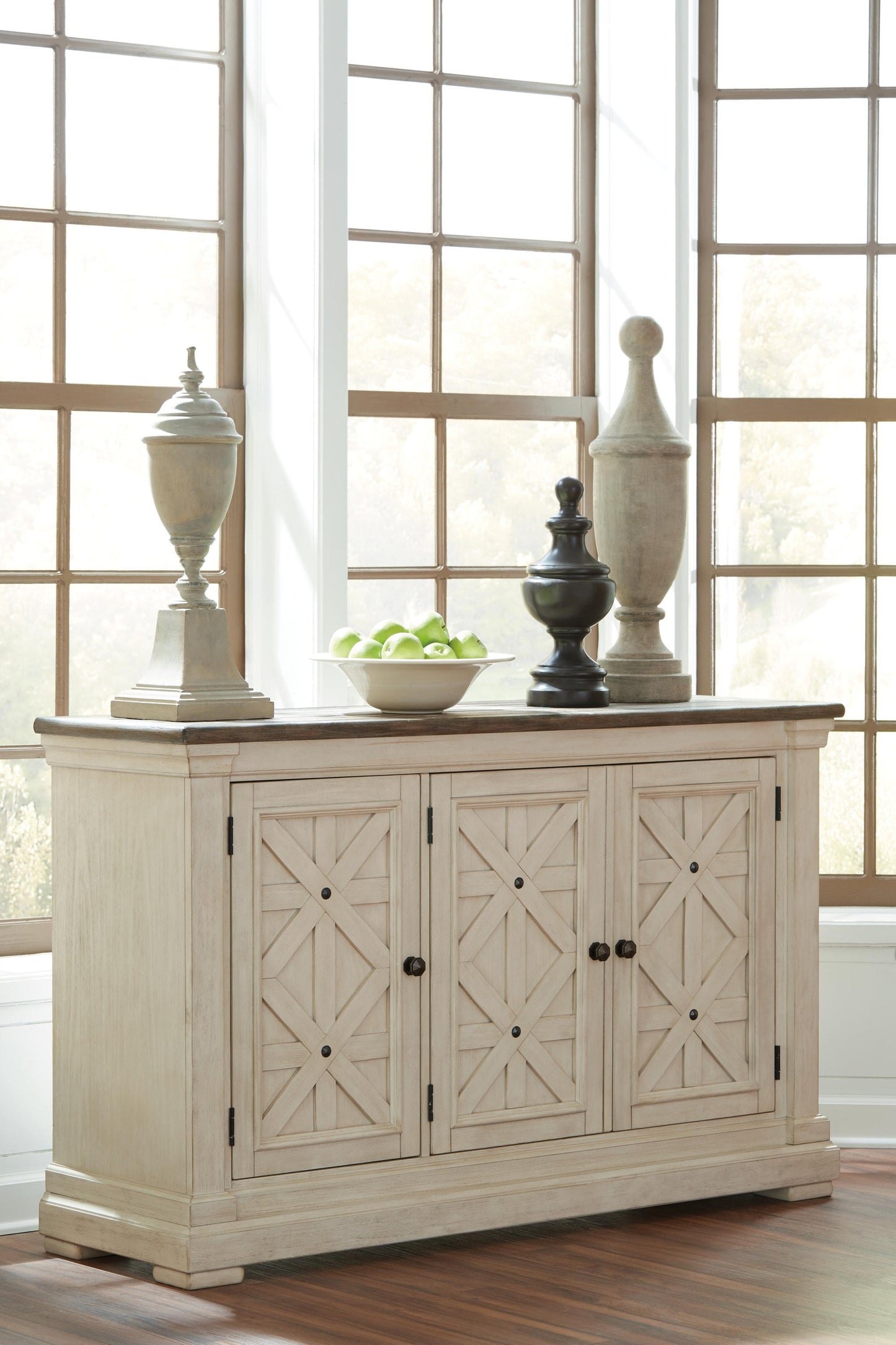 The Bolanburg Dining Room Server features a light wood finish and geometric patterns. Positioned by large windows, it is adorned with three decorative sculptures and a bowl of green apples.