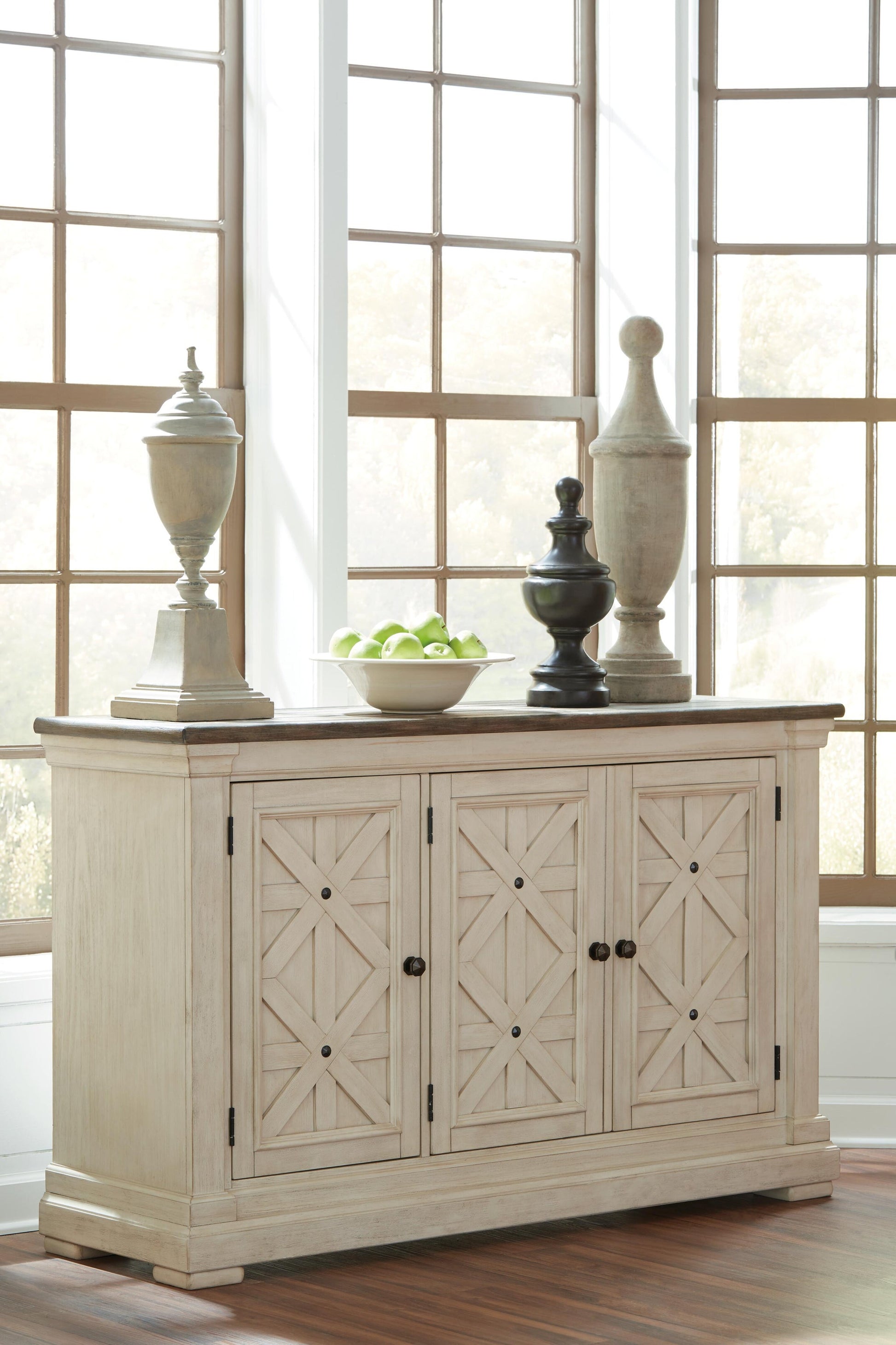 The Bolanburg Dining Room Server features a light wood finish and geometric patterns. Positioned by large windows, it is adorned with three decorative sculptures and a bowl of green apples.