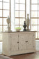 The Bolanburg Dining Room Server features a light wood finish and geometric patterns. Positioned by large windows, it is adorned with three decorative sculptures and a bowl of green apples.