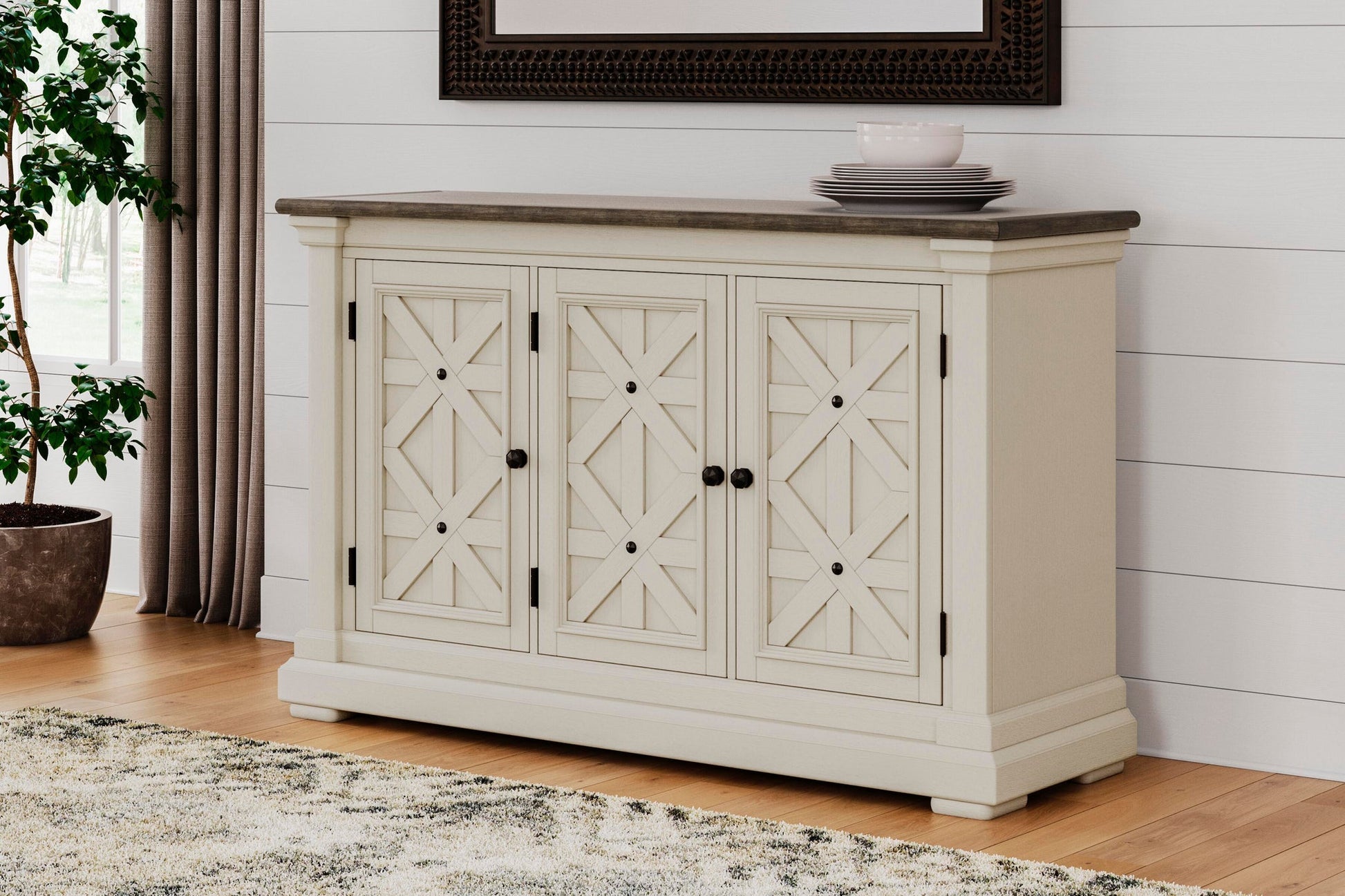 The Bolanburg Dining Room Server, featuring a cream finish with three paneled doors and black knobs, stands against a white shiplap wall, topped with stacked plates, next to a plant and rug.