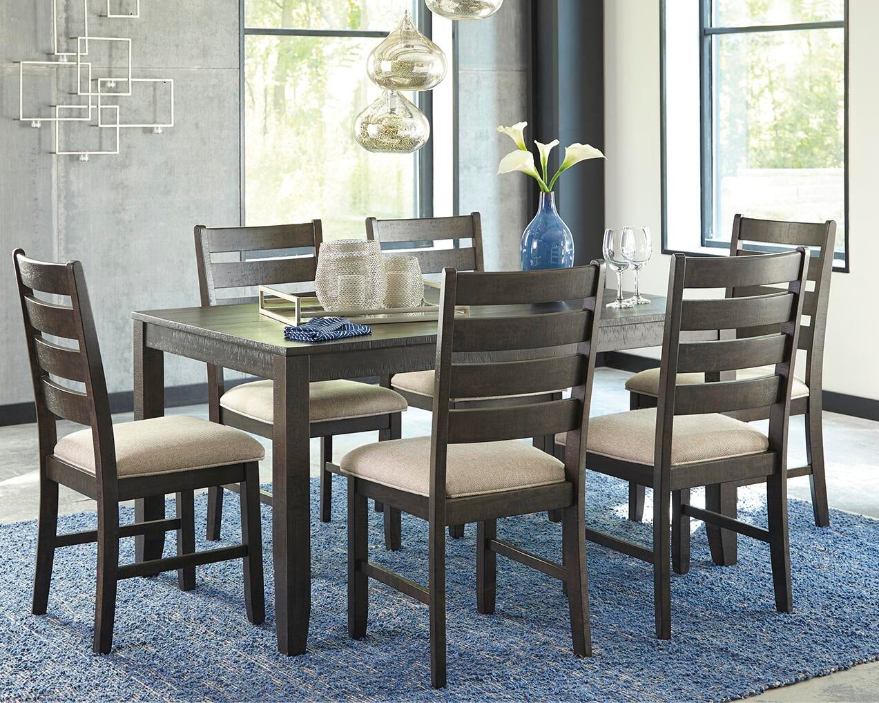 A modern dining room showcases the Rokane Dining Room Table Set (7/CN) with six beige upholstered chairs, a blue rug, pendant lighting, and a blue vase filled with white flowers.
