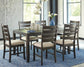 A modern dining room showcases the Rokane Dining Room Table Set (7/CN) with six beige upholstered chairs, a blue rug, pendant lighting, and a blue vase filled with white flowers.