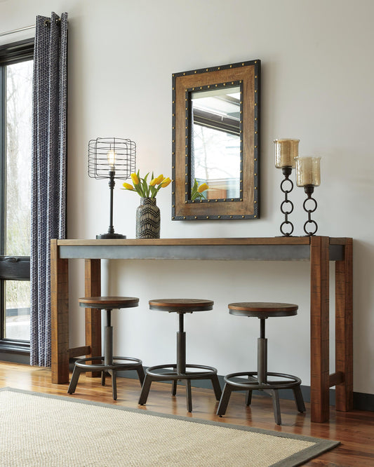 The Torjin Long Counter Table is paired with three stools, a lamp, yellow tulips in a vase, two candle holders, and a framed mirror on the wall in a modern room.