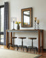 The Torjin Long Counter Table with three stools, a lamp, a vase of yellow tulips, two candle holders, and a framed mirror creates a cozy scene against a white wall.