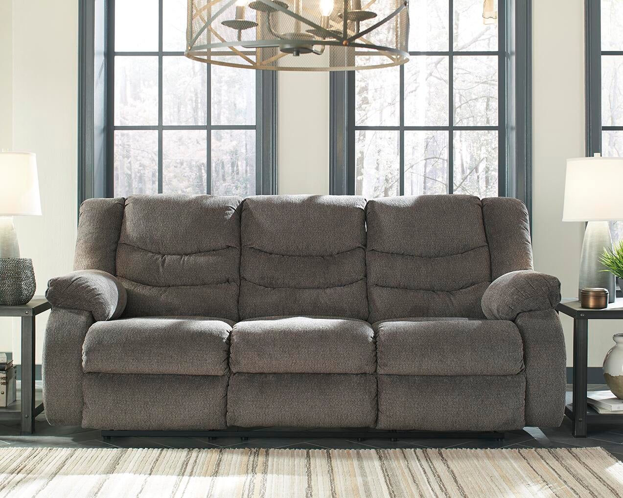 The Tulen Reclining Sofa, a gray three-seater with plush chenille cushions, is shown in a living room between two lamp-topped side tables and large windows in the background.