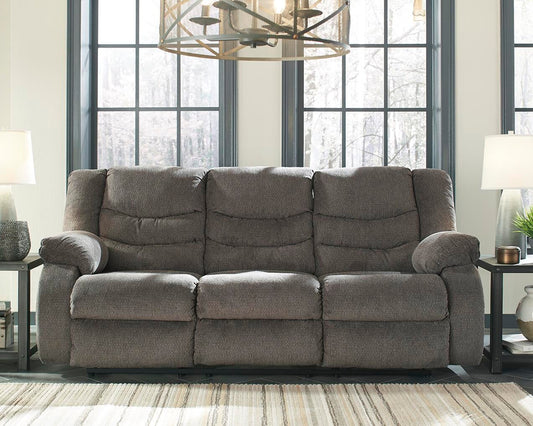 The Tulen Reclining Sofa, a gray three-seater with plush chenille cushions, is shown in a living room between two lamp-topped side tables and large windows in the background.