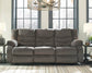 The Tulen Reclining Sofa, a gray three-seater with plush chenille cushions, is shown in a living room between two lamp-topped side tables and large windows in the background.