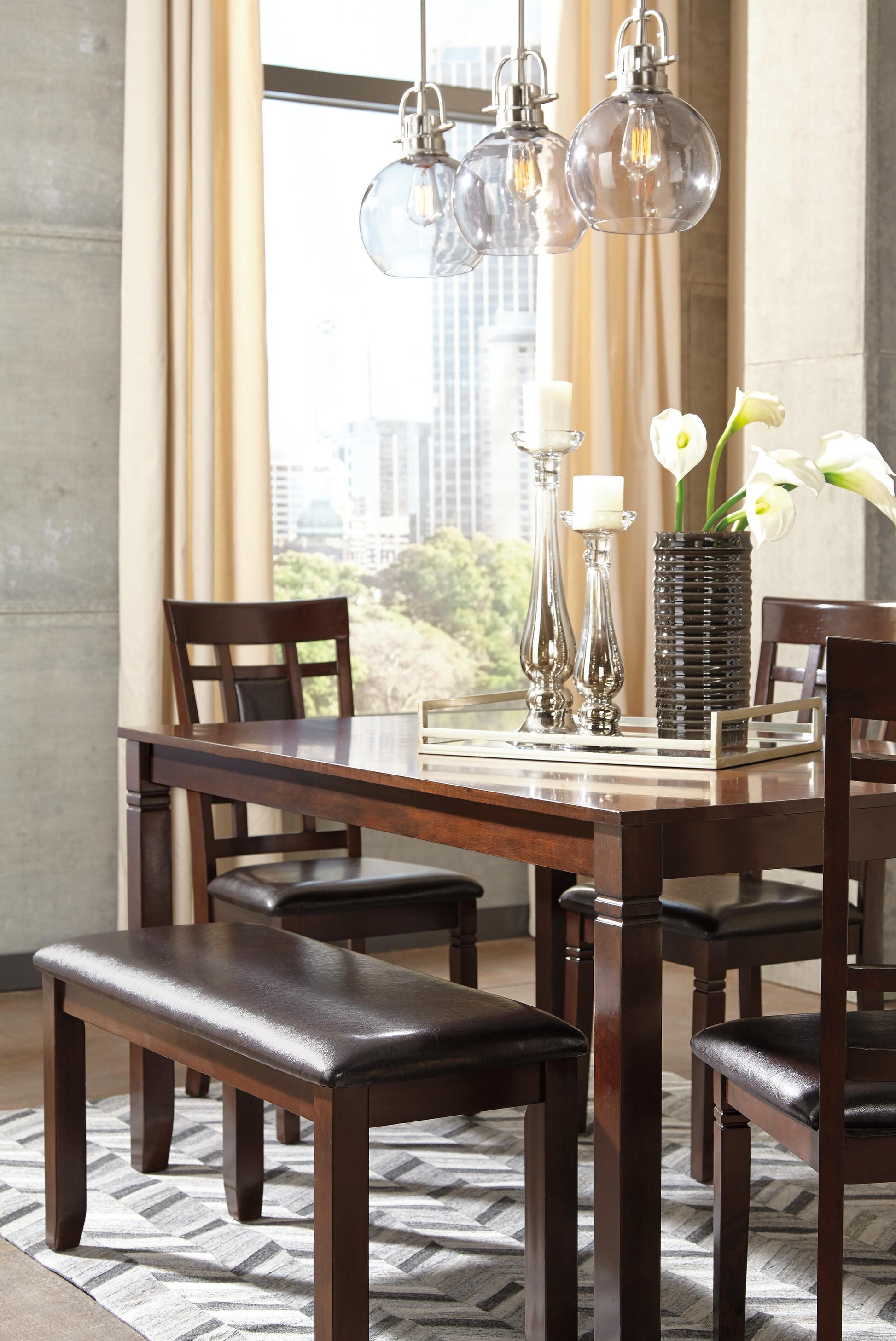 A modern dining room features the Bennox Dining Room Table Set (6/CN) with upholstered chairs, matching bench, decorative candles, white flowers in a vase, and glass pendant lights, framed by large windows showcasing a stunning city view.