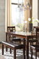 A modern dining room features the Bennox Dining Room Table Set (6/CN) with upholstered chairs, matching bench, decorative candles, white flowers in a vase, and glass pendant lights, framed by large windows showcasing a stunning city view.