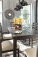 A modern dining room highlights the Rokane Dining Room Table Set (7/CN), surrounded by large windows, gray walls, glass jars with lemons, elegant glassware, and stylish pendant lights.