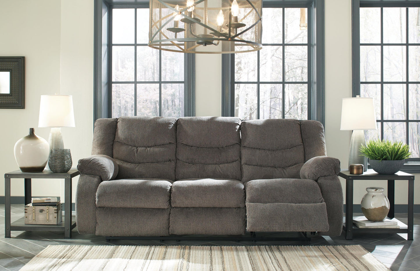 The Tulen Reclining Sofa in soft gray chenille sits between two side tables with lamps and decor, in front of large windows brightening the living room.