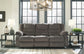 A Tulen Reclining Sofa in gray chenille fabric is centered between two matching side tables with lamps before large windows. Decorative accents and a neutral rug complete the living room setup.