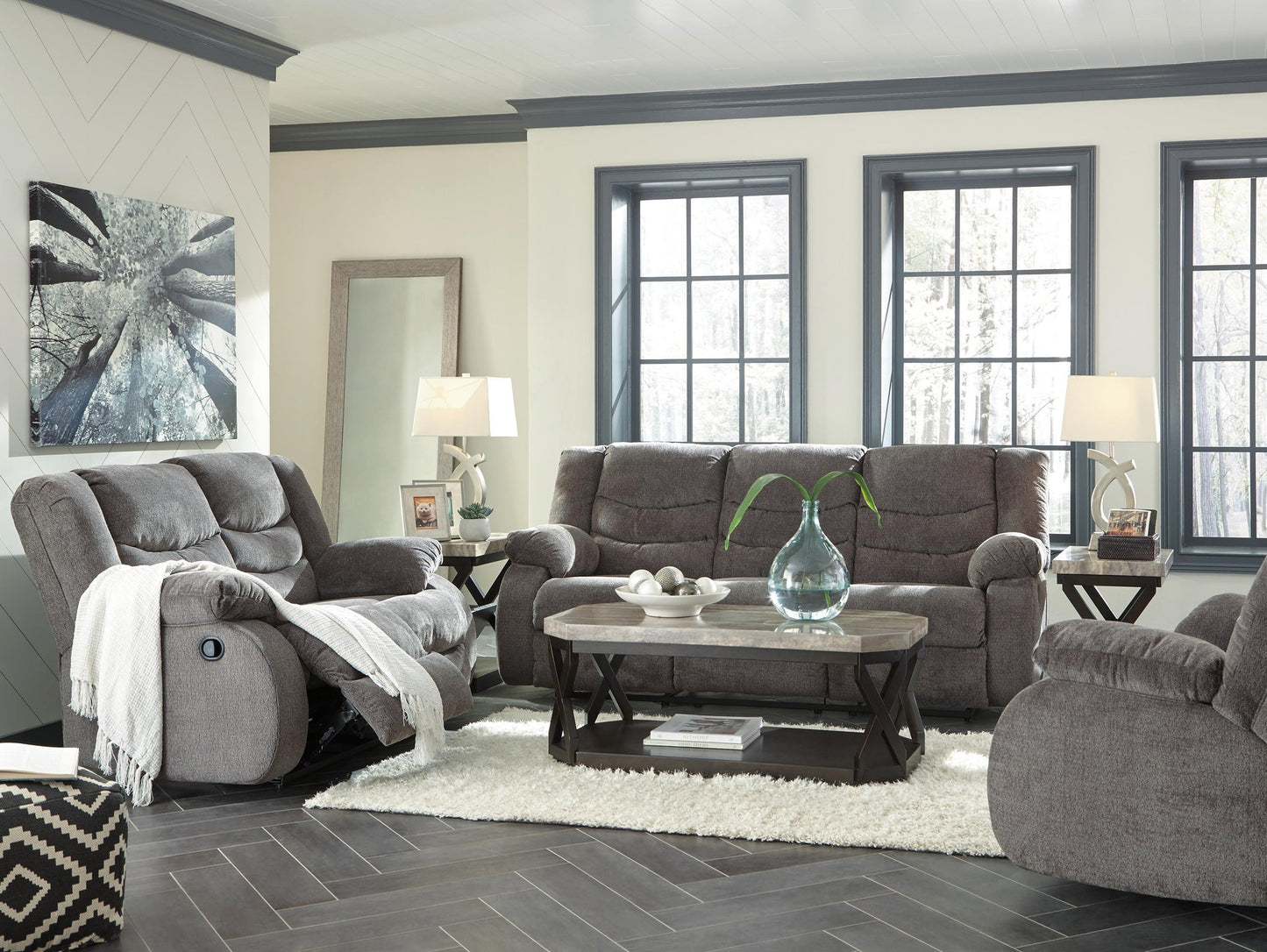 A living room featuring the Tulen Reclining Sofa in gray chenille fabric, a wooden coffee table with a glass vase, two lamps, a rug, a wall mirror, and large windows letting in natural light.