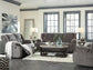 A living room featuring the Tulen Reclining Sofa in gray chenille fabric, a wooden coffee table with a glass vase, two lamps, a rug, a wall mirror, and large windows letting in natural light.