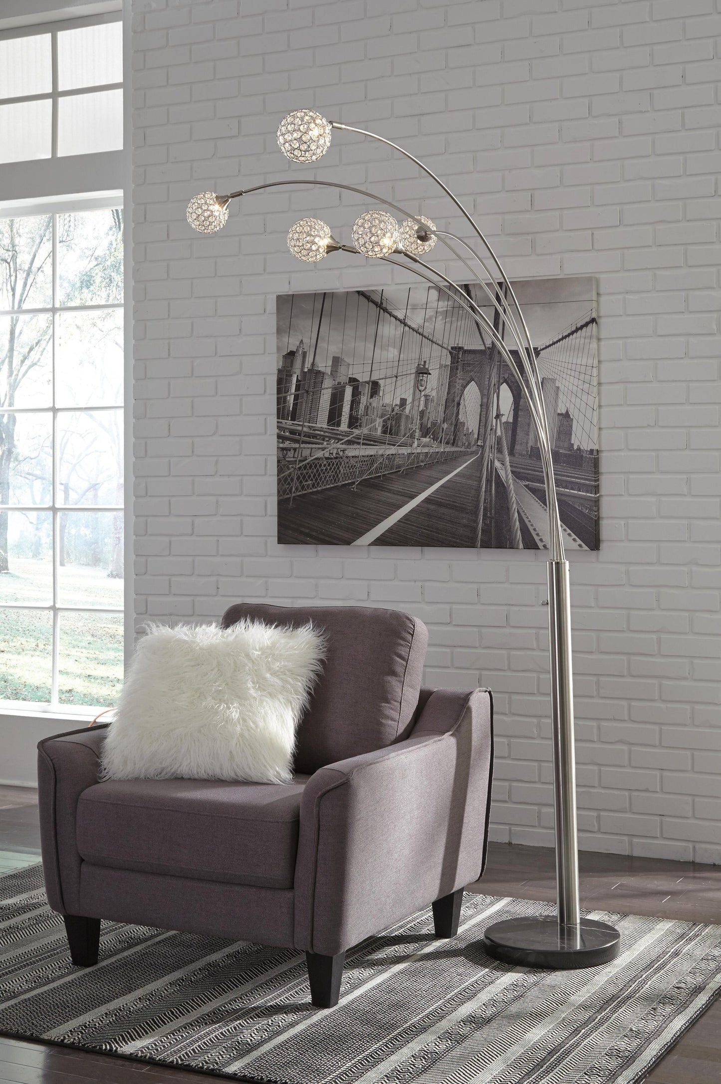 A gray armchair with a white pillow sits beside the Winter Metal Arc Lamp and a black-and-white bridge photo on a white brick wall in a bright room.