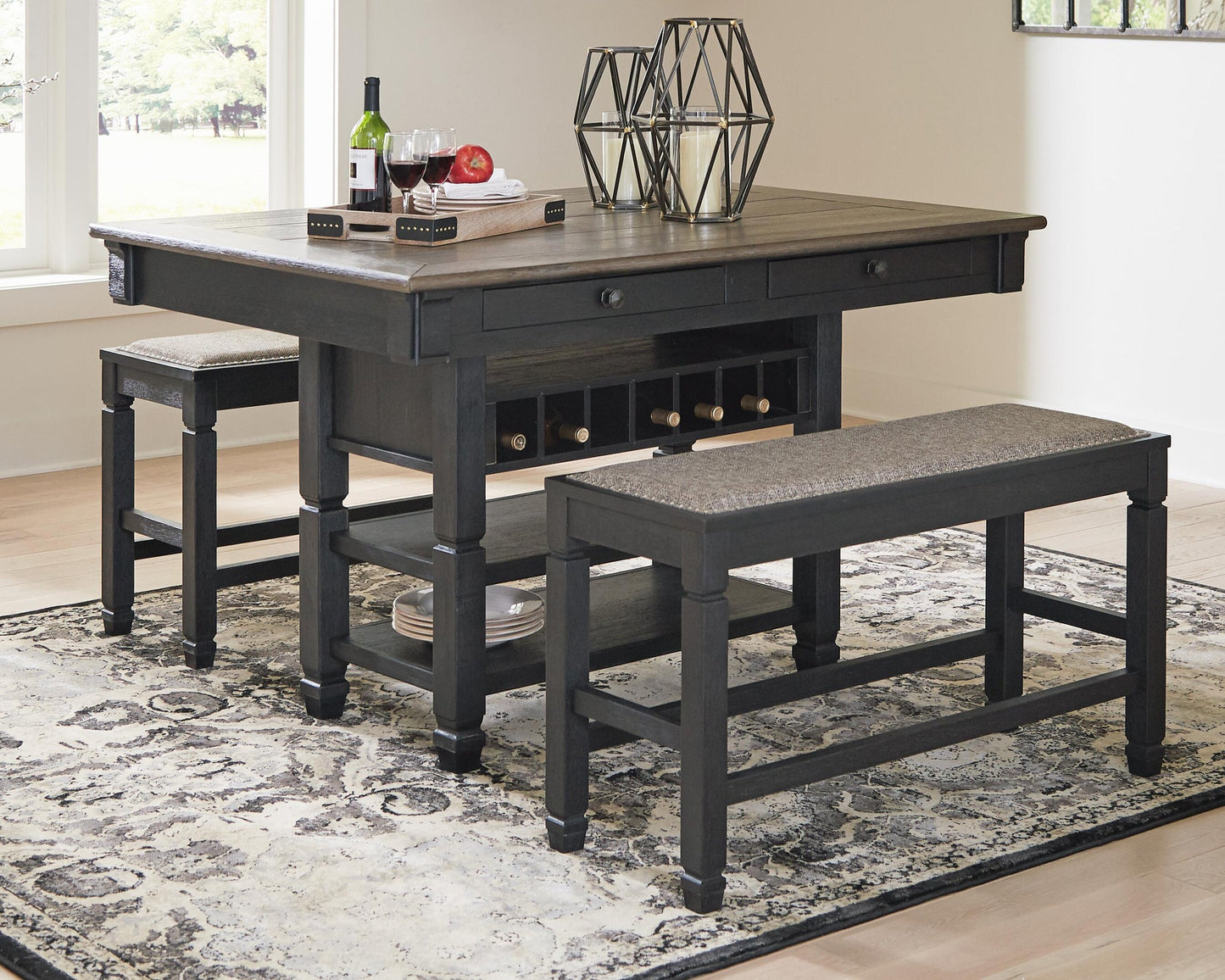 The Tyler Creek RECT Dining Room Counter Table with wine storage and two benches, in dark wood, is styled with decor on a patterned rug in a bright dining nook for cozy urban farmhouse charm.