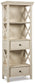 The Bolanburg Display Cabinet features an antiqued white finish, open sides with crisscross details, two shelves, and two drawers with round knobs—a charming cottage-style addition to your home.