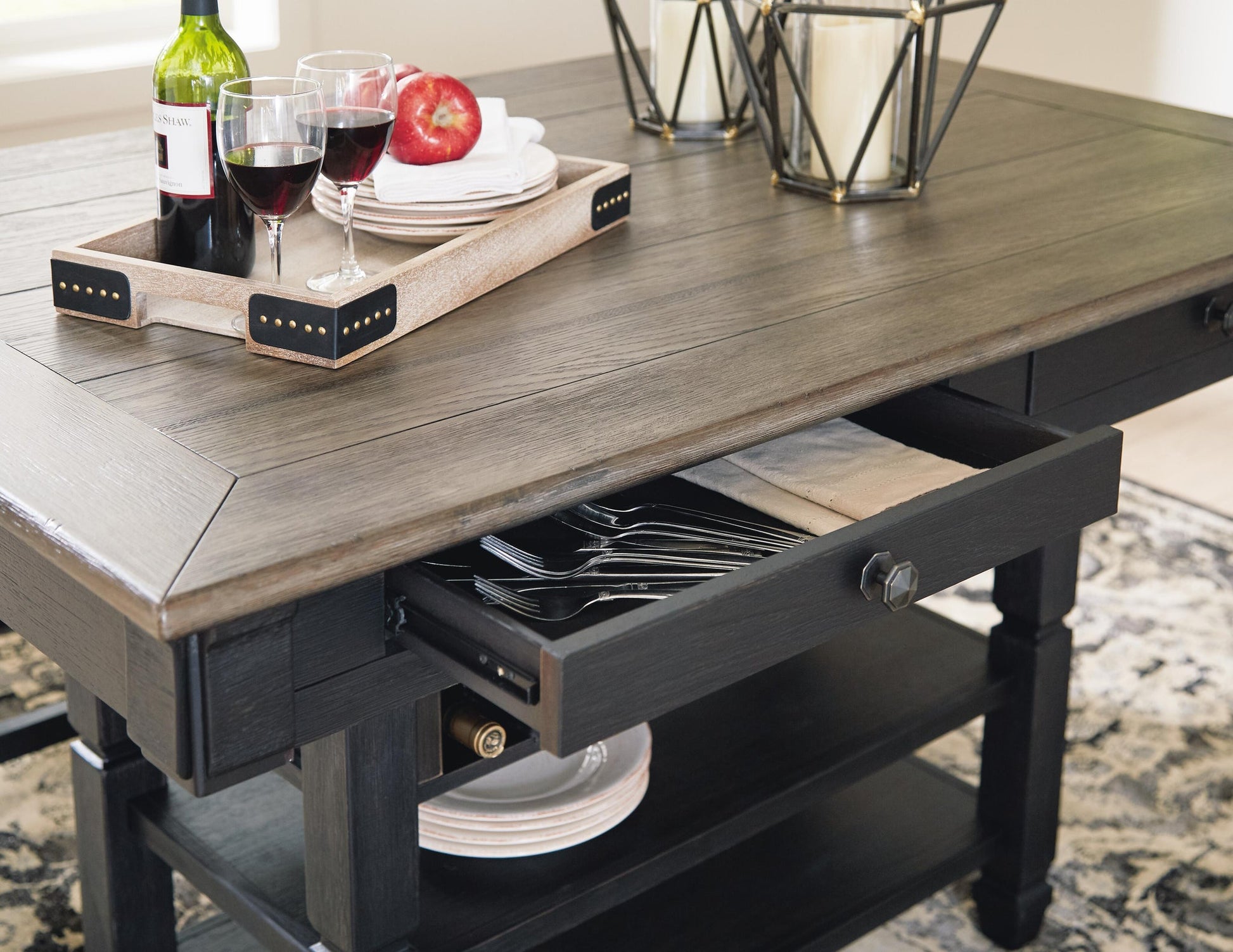 The Tyler Creek RECT Dining Room Counter Table showcases urban farmhouse style, with a partially open cutlery drawer, a tray holding wine glasses and a bottle, candles, and a plate with an apple on top.