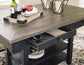 The Tyler Creek RECT Dining Room Counter Table showcases urban farmhouse style, with a partially open cutlery drawer, a tray holding wine glasses and a bottle, candles, and a plate with an apple on top.