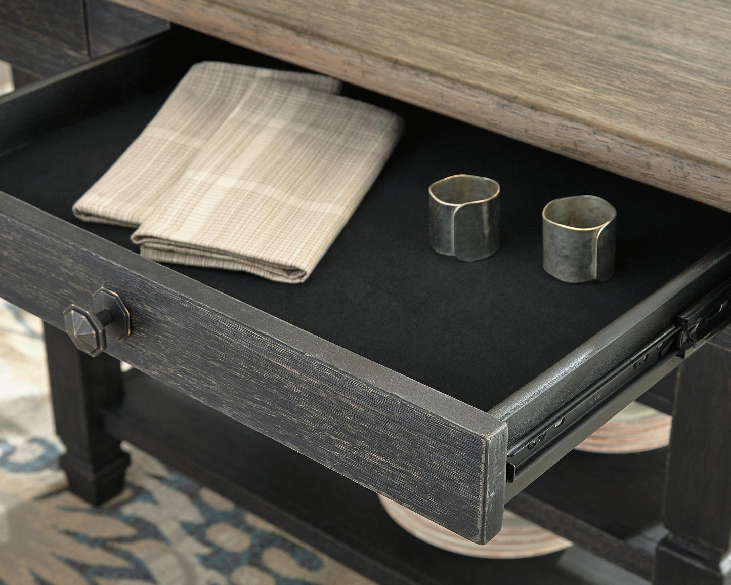 An open drawer in the Tyler Creek RECT Dining Room Counter Table holds two folded plaid napkins and two metal napkin rings on a black liner, bringing urban farmhouse style to your space.